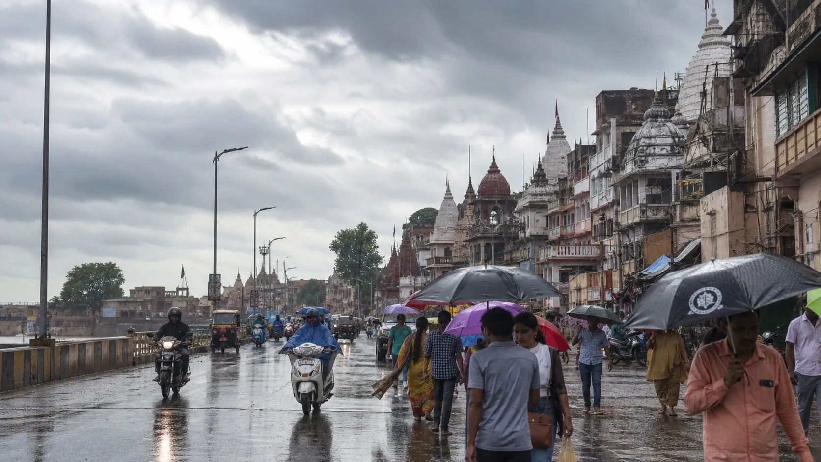 Cloudy monsoon sky above a rain-damp Varanasi road with commuters carrying umbrellas