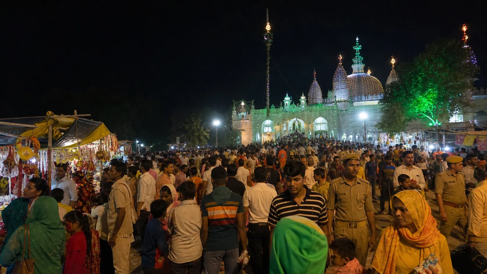 Families and pilgrims walking past illuminated stalls at a dargah fair near Pindra