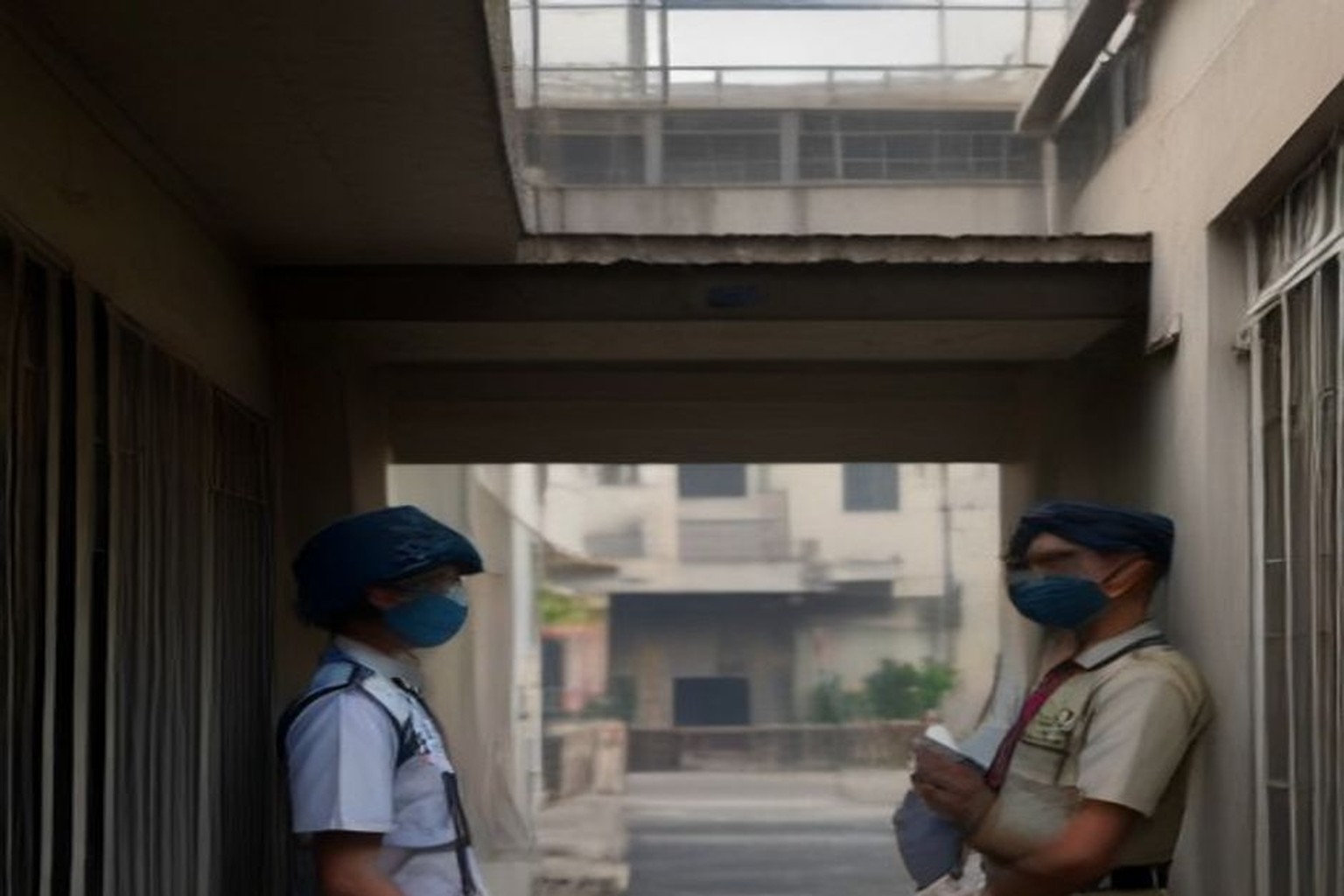 Two masked officers in caps stand under a concrete walkway between residential buildings, one holding papers, in a bright Gurugram-style colony alley.
