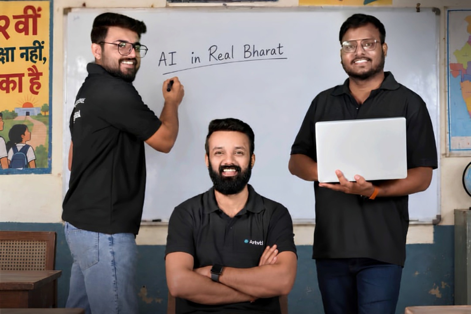 Three men in matching black polo shirts in a classroom, one writing on a whiteboard that reads AI in Real Bharat